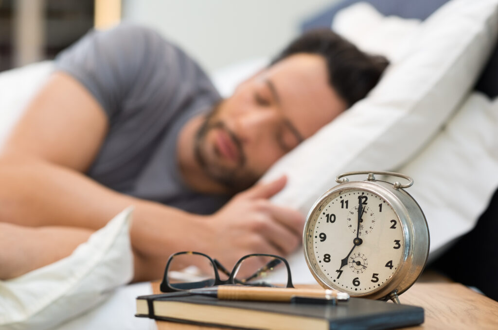Man sleeping with glasses and an alarm clock on his bedside table.