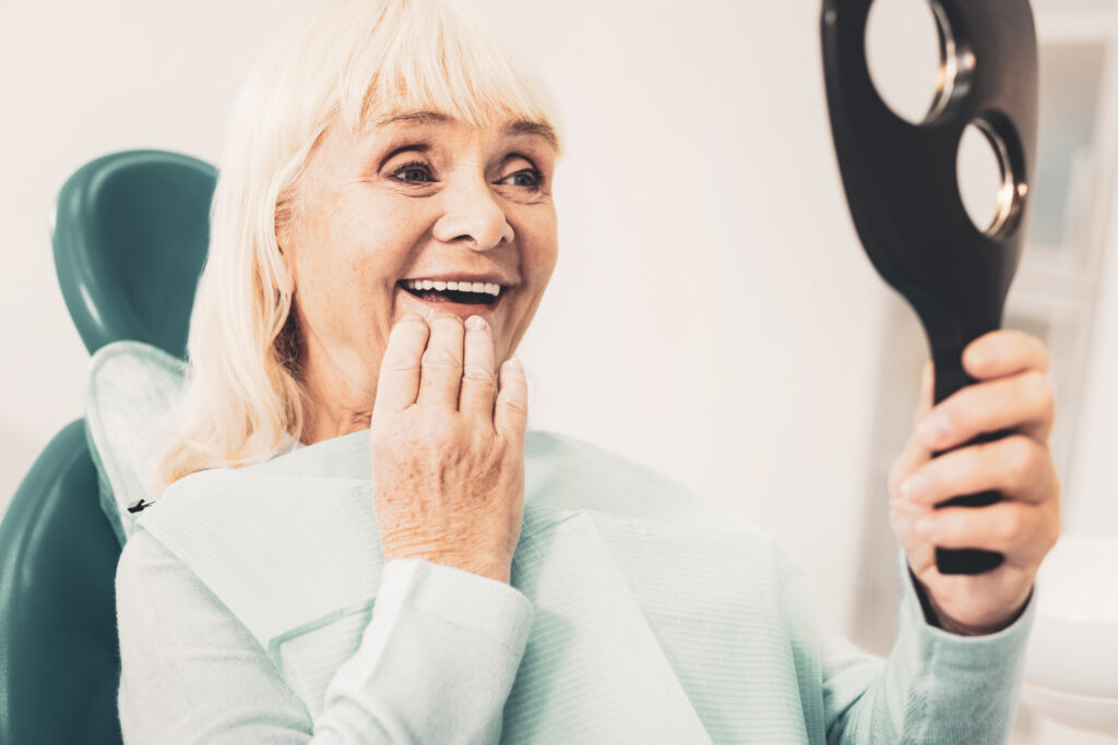 A smiling woman with dentures look into a handheld mirror