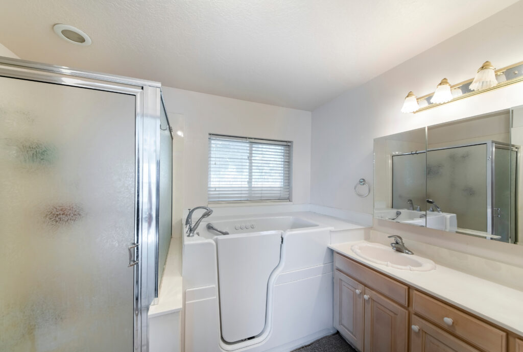 Walk-in bathtub with elderly and handicapped accessibility. There is a vanity sink with mirrors on the right and shower stall with frosted glass and aluminum frames.