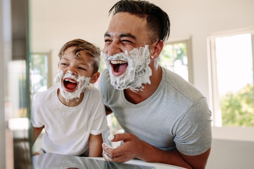 Man and little boy with shaving foam on their faces looking into the bathroom mirror and laughing.