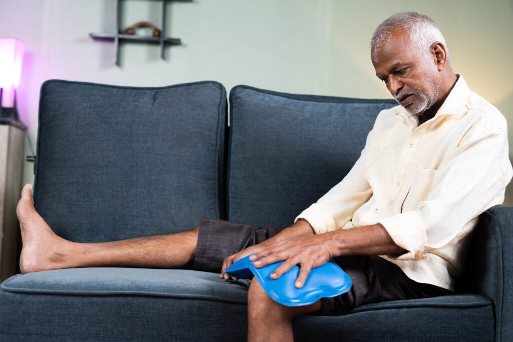 senior man using Hot water massage bag for knee joint pain relief while sitting on sofa at home - concept of natural hot water treatment, muscle relaxtion and osteoarthritiss